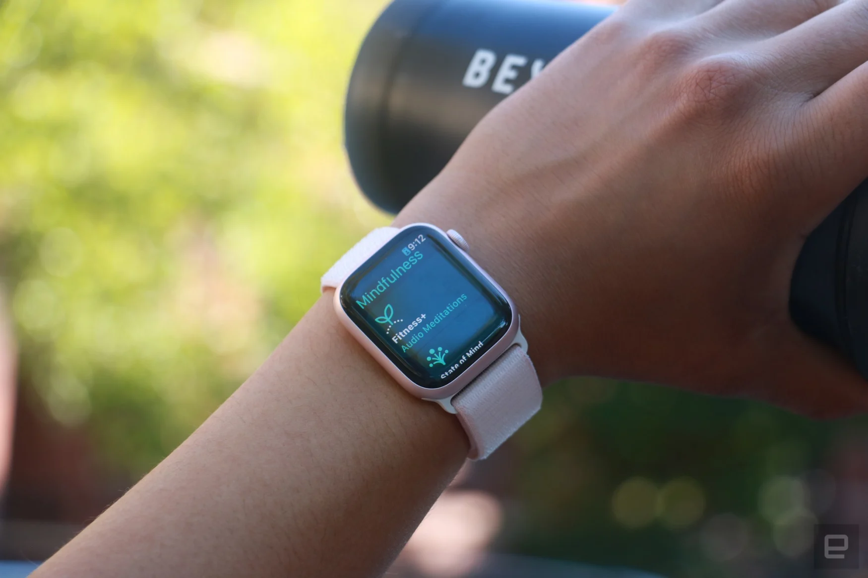 The Apple Watch Series 9 on a person's wrist, with the hand holding a black water bottle in the background. The screen shows the Mindfulness app.