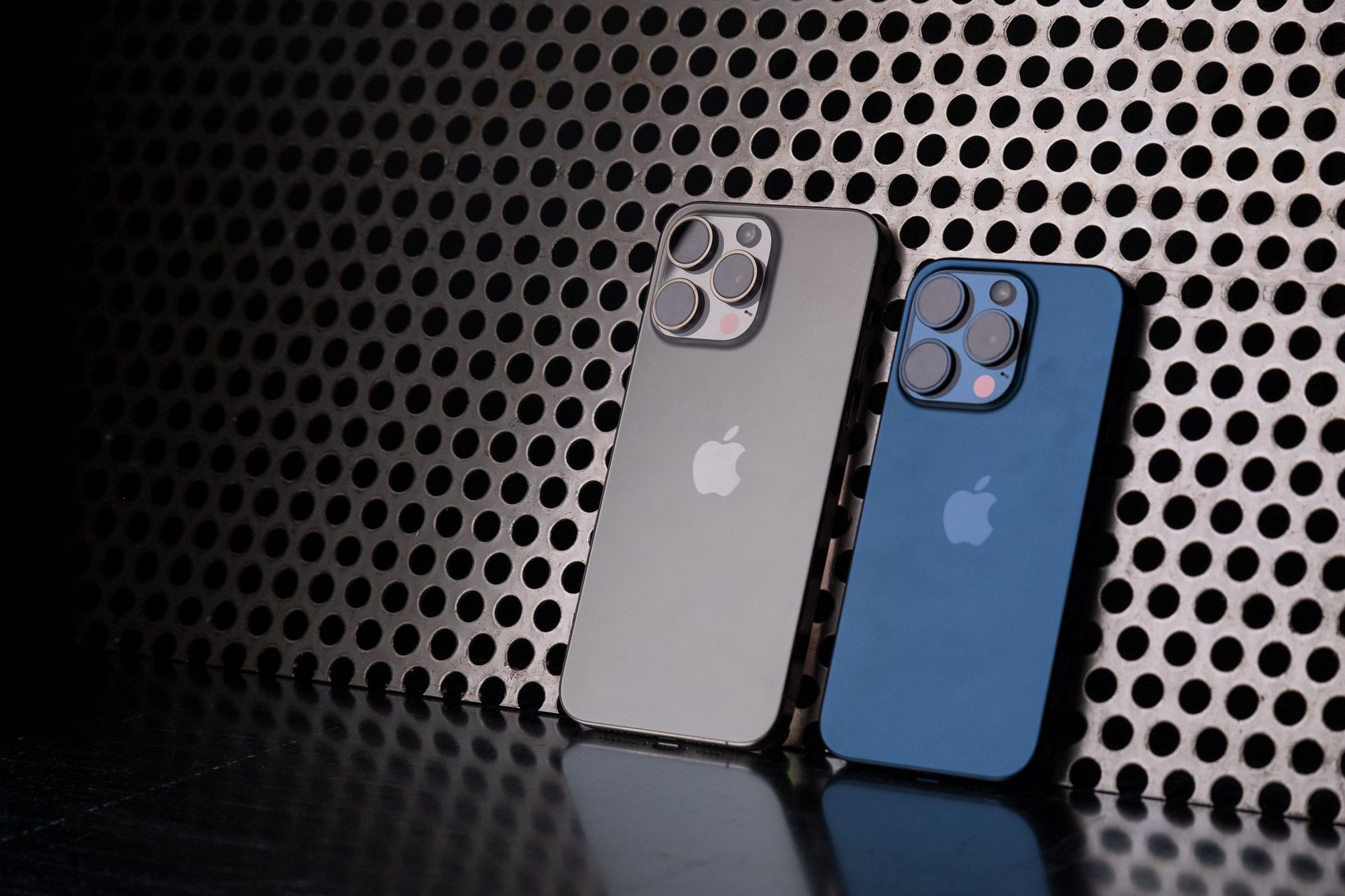 Blue iPhone 15 Pro and natural titanium iPhone 15 Pro Max leaning against a perforated metal background.